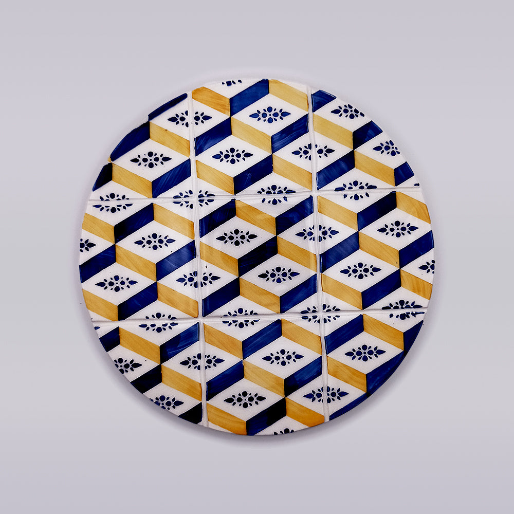 The Algarve Ceramic Kitchen Trivet by Tejo Shop features a round ceramic plate with a geometric pattern, showcasing blue, yellow, and white hexagons adorned with small black floral designs on a plain light grey background. This hand-crafted design creates an optical illusion of three-dimensional cubes, making it perfect as an artisanal trivet.