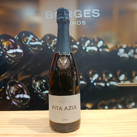 A bottle of Fita Azul Attitude Reserve Brut by Borges Wines stands on a light wooden surface. Blurred wine bottles fill the background, with "BORGES" partially visible. Fine bubbles hint at its refined character.