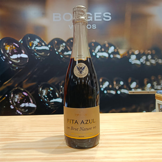 A bottle of Borges Wines Fita Azul Brut Nature Sparkling Wine Reserve rests on a wooden surface, its gold label and foil shining, ideal for Gouveio lovers in a wine shop or cellar with rows of bottles blurred behind.