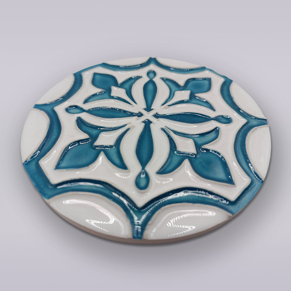 The Portuguese Flor-de-Lis Tile – Atlantic Edition by Miradouro Ceramics is a round ceramic tile with a raised teal and white floral-cross motif on a light background, ideal as a decorative accent.