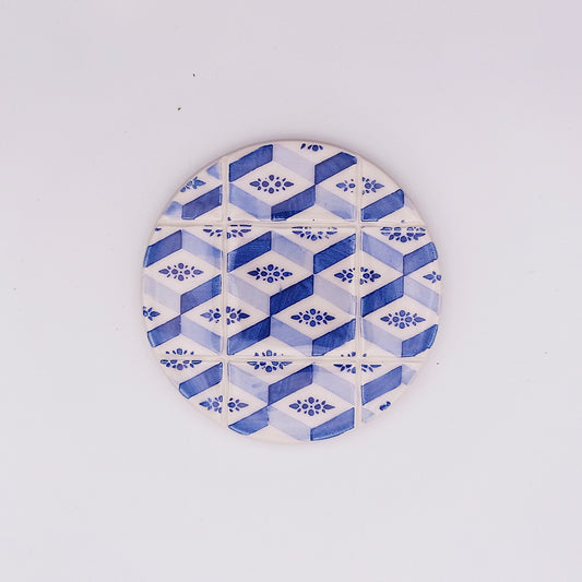 A round ceramic coaster with a geometric pattern of blue and white hexagons, each containing a small floral design at the center, arranged in a grid formation against a white background, exudes an artisanal charm, reminiscent of the hand-made Estoril Ceramic Tile Cup Pad by Tejo Shop.