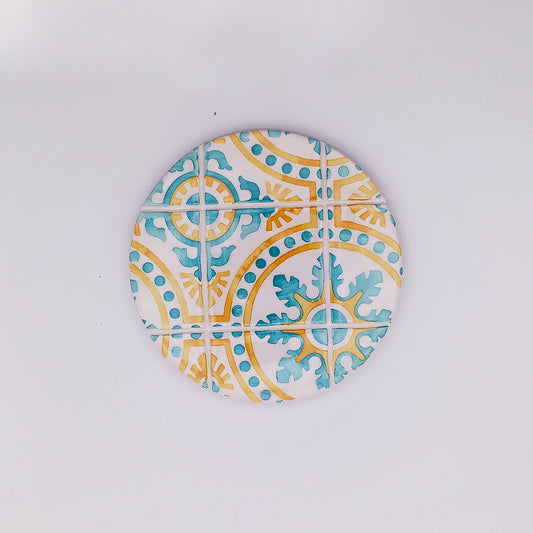 A round, ceramic coaster featuring a decorative pattern of blue and yellow geometric and floral-inspired designs. Reminiscent of a hand-painted ceramic tile, the pattern consists of intersecting lines and dots, creating an eye-catching, symmetrical arrangement. The background is white. This is the Fatima Ceramic Cup Pad by Tejo Shop.