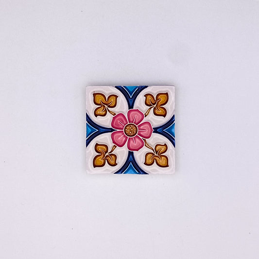 Square decorative hand-painted Tejo Shop small ceramic tile with a central pink flower design, surrounded by blue and gold patterns on a white background.