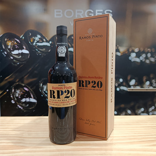 A bottle of Ramos Pinto 20 Year Old Tawny Porto - Quinta do Bom Retiro stands on a wooden surface beside its matching tall brown box, with blurred wine bottles visible in the background.