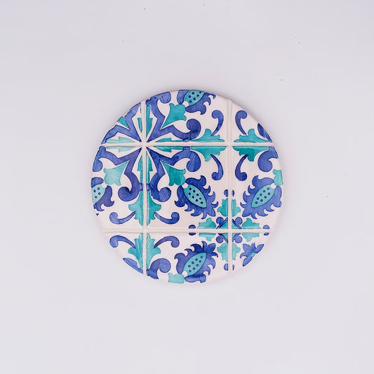 A circular ceramic coaster, hand-made and hand-painted with a traditional intricate blue and green floral pattern on a white background, reminiscent of Portuguese azulejo tiles. The design features symmetrical floral and geometric elements, perfect to protect your counters. Meet the Albufeira Ceramic Cup Pad by Tejo Shop.