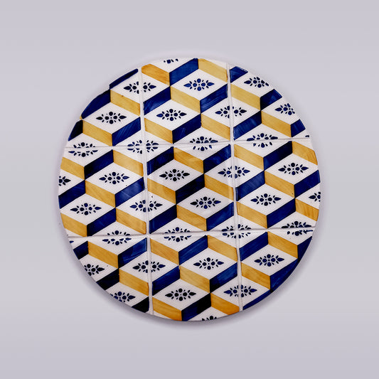 The Algarve Ceramic Kitchen Trivet by Tejo Shop features a round ceramic plate with a geometric pattern, showcasing blue, yellow, and white hexagons adorned with small black floral designs on a plain light grey background. This hand-crafted design creates an optical illusion of three-dimensional cubes, making it perfect as an artisanal trivet.