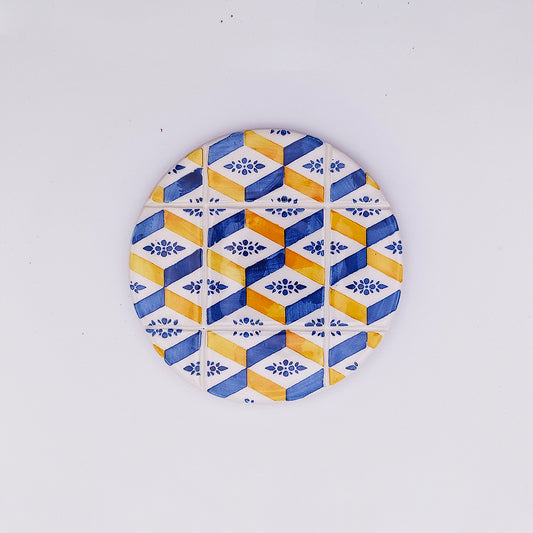 A round Algarve Ceramic Tile Cup Pad with a geometric pattern of yellow, blue, and white hexagons and diamonds. The pattern includes floral motifs within the hexagons, creating a visually appealing and decorative design. This hand-made quality ceramic tile coaster from Tejo Shop is centered on a plain white background.
