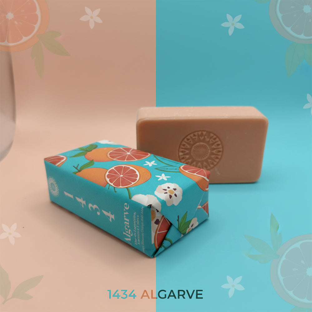 A bar of Algarve 1434 – Edge of the Known World soap by 100ml, in a colorful fruit-patterned wrapper, sits on a peach and teal background with citrus and floral graphics. Handcrafted in Portugal.