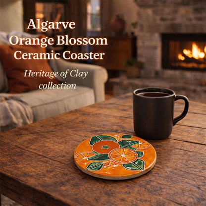 The Miradouro Ceramics Algarve Orange Blossom Ceramic Coaster, hand-painted with a vibrant orange blossom design, sits by a black coffee mug on a wooden table. A fireplace and cozy living room glow warmly in the background.