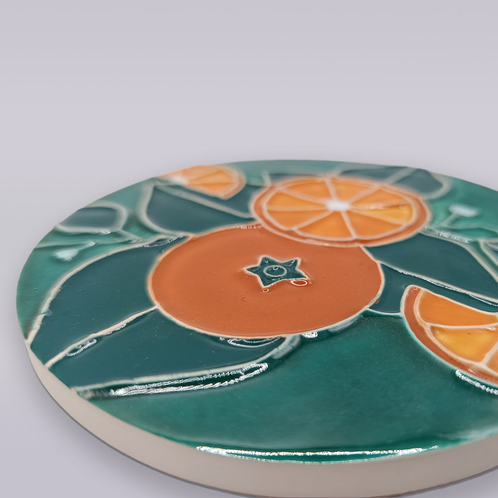 The Algarve Citrus Grove Ceramic Coaster by Miradouro Ceramics features a glossy round surface with an artistic orange and green leaf design, capturing the vibrant charm of hand-painted Algarve citrus groves.