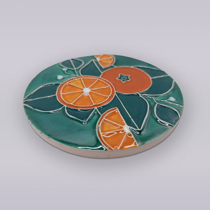 The Algarve Citrus Grove Ceramic Coaster by Miradouro Ceramics features a round Portuguese design with orange slices and green leaves, adding hand-painted Algarve charm and citrus grove decor to any setting.