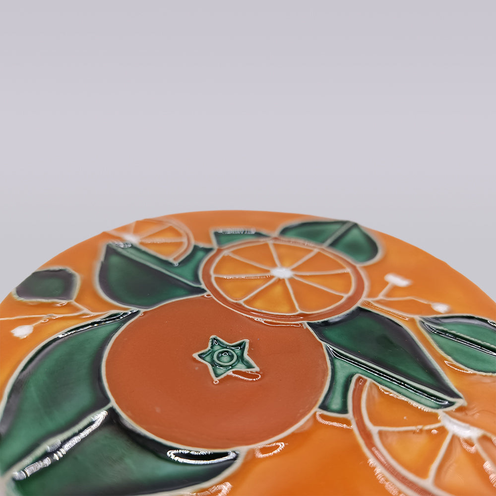 The Algarve Orange Blossom Ceramic Coaster by Miradouro Ceramics features a hand-painted orange fruit and green leaves design with a glossy finish, shown close-up against a light gray background.