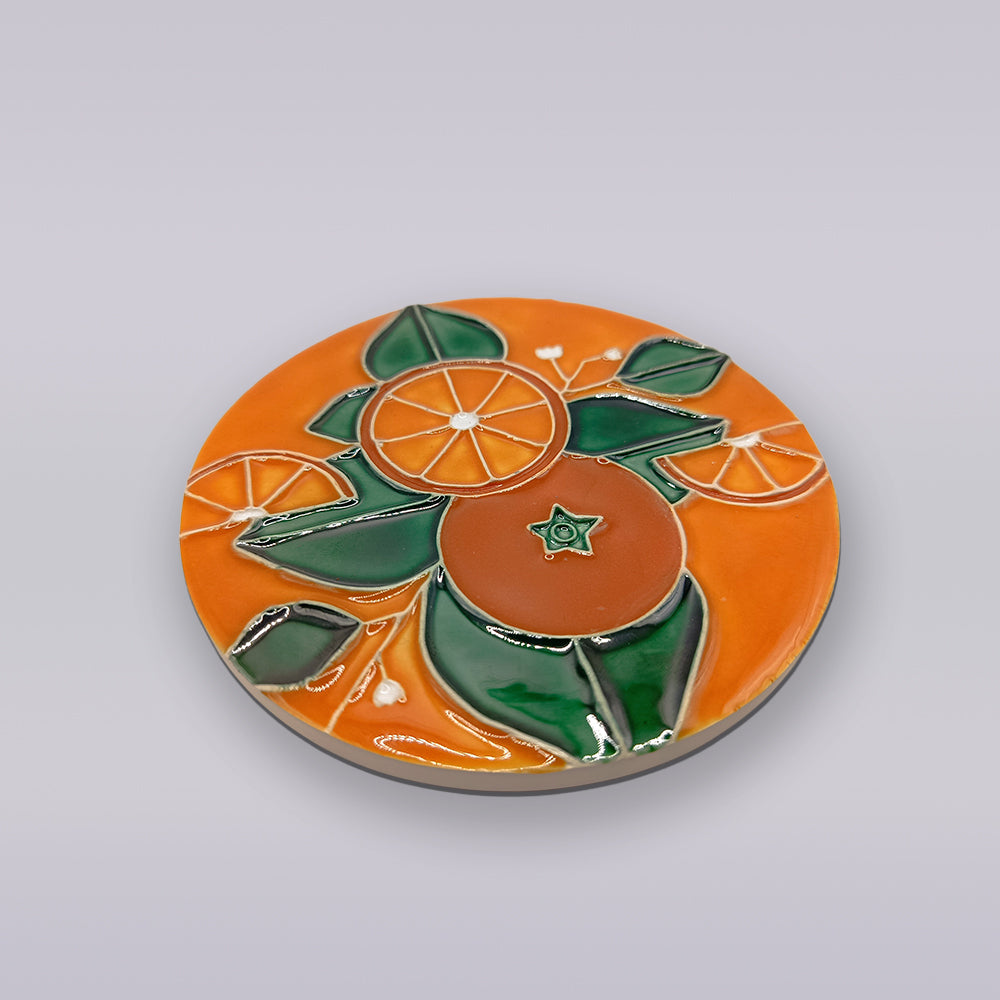 The Algarve Orange Blossom Ceramic Coaster by Miradouro Ceramics features a glossy orange surface with a raised orange fruit, slices, and green leaves, inspired by Algarve coaster designs and shown on a plain light gray background.