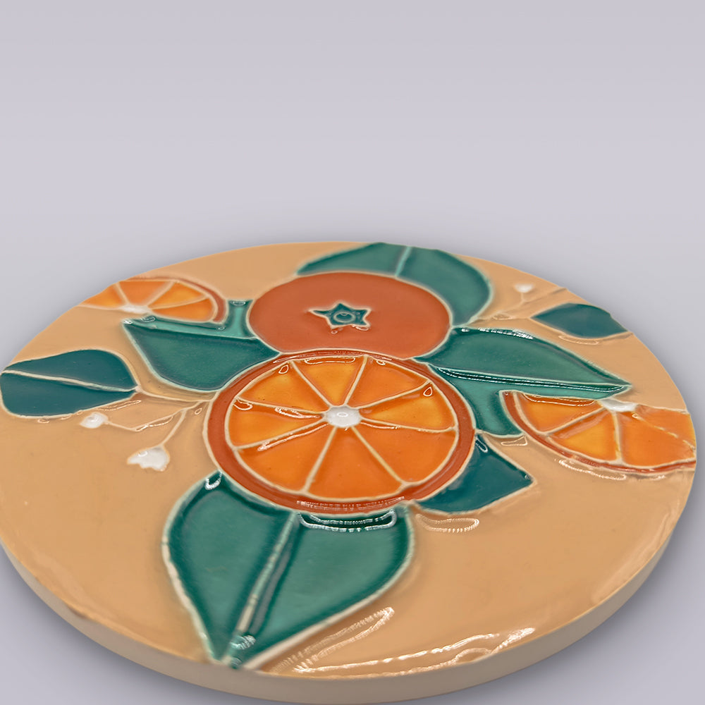 The Miradouro Ceramics Algarve Sandstone Orange Ceramic Coaster features embossed orange citrus slices and green leaves on a beige background, finished with a glossy sheen.