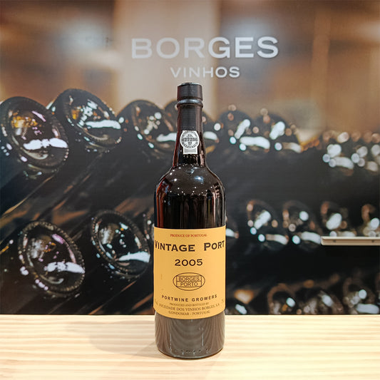 A bottle of Borges Vintage 2005 by Borges Wines stands on a light wooden surface, with stacked wine bottles and the blurred words “Borges Vinhos” in the background.