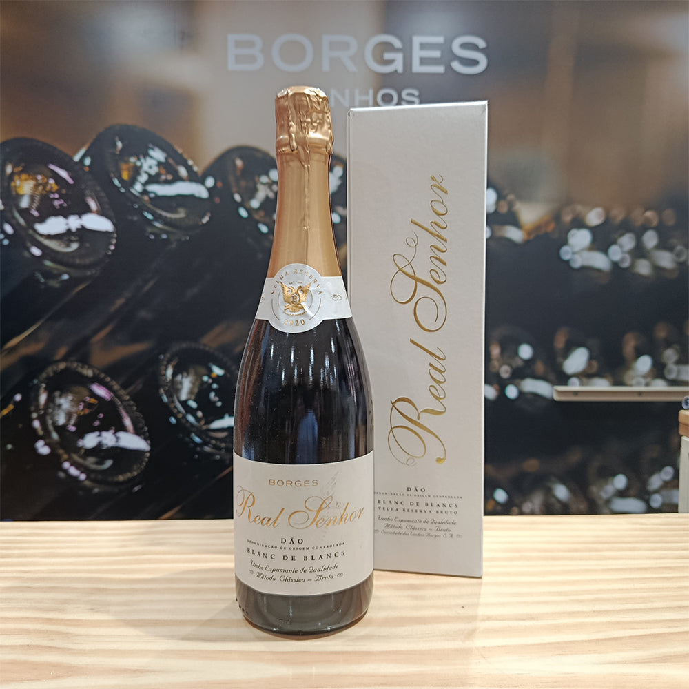 A bottle of Borges Wines Real Senhor Blanc de Blancs Old Reserv Brut stands on a wooden surface beside its elegant white and gold box, with blurred wine bottles in the background, evoking its refined, fruity aroma.
