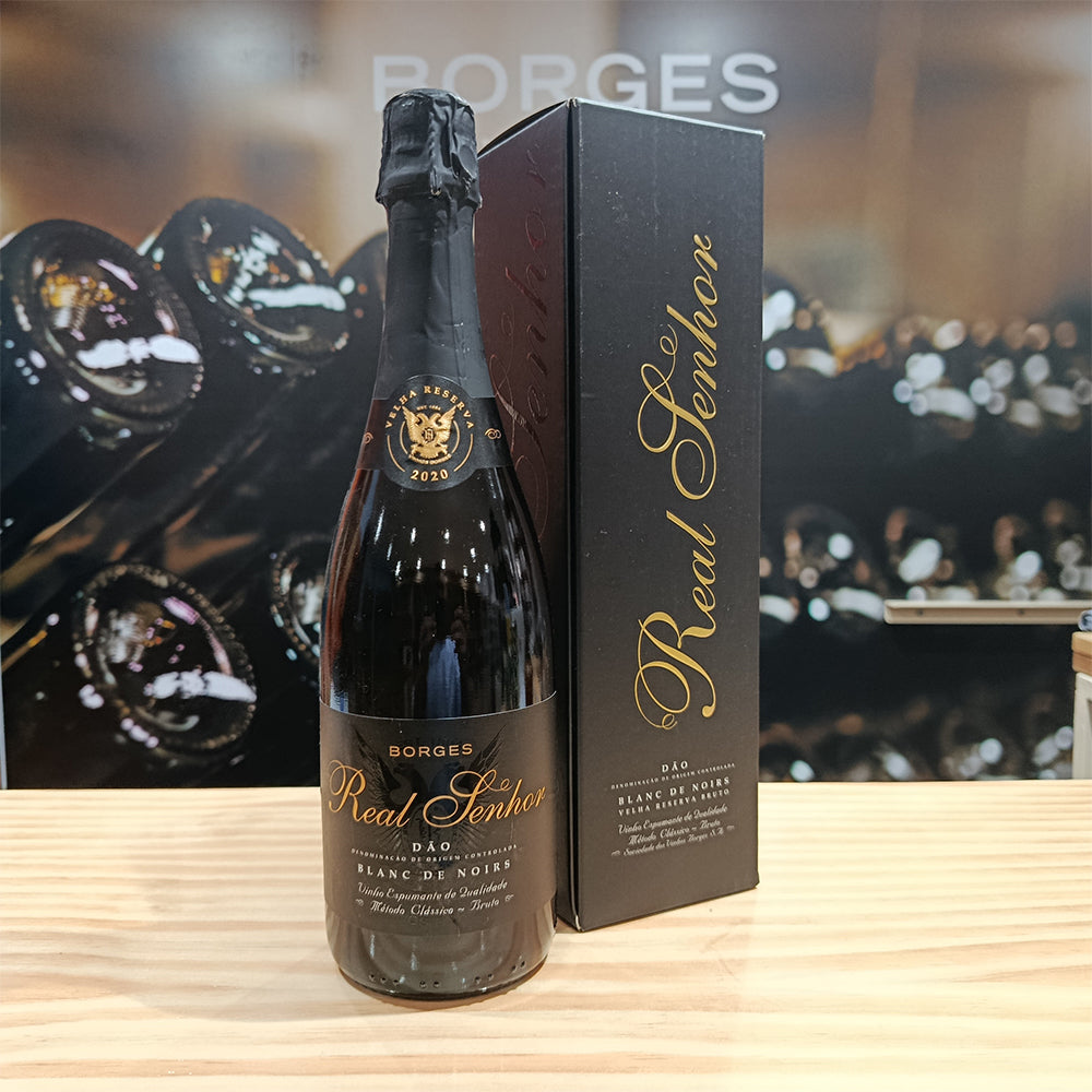 A bottle of Borges Wines Real Senhor Blanc de Noirs Old Reserv Brut stands next to its black and gold box on a wooden surface, with blurred bottles—including Borges Real Senhor Blanc de Blancs—visible in the background.