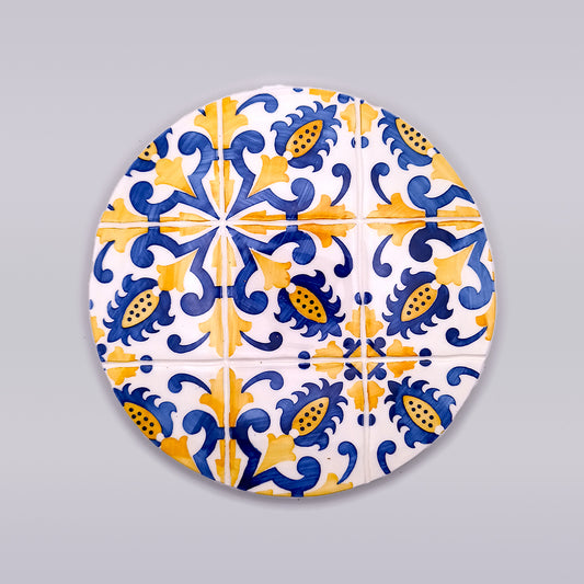 A circular, hand-painted ceramic trivet featuring a symmetrical design with blue and yellow floral and geometric patterns on a white background. The intricate motifs are reminiscent of traditional Mediterranean or Spanish tile work. The Braga Ceramic Trivets for Hot Dishes by Tejo Shop is set against a plain gray backdrop.