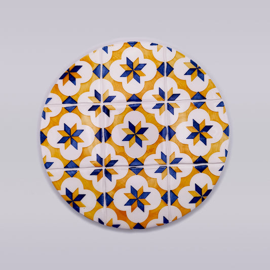 Image of a round, Cascais Ceramic Hot Plate Trivet with a geometric pattern of star shapes and flowers in blue, white, and yellow. The hand-painted ceramic tile design is reminiscent of traditional tile motifs. The background is a plain, light gray. This durable and elegant trivet from Tejo Shop adds charm to any setting.