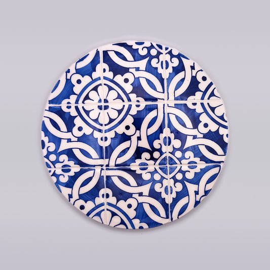 A round blue and white decorative plate with an intricate geometric pattern, hand-painted for a unique touch. The design features interlocking shapes resembling flowers and leaves, creating a mosaic-like appearance. This exquisite piece, reminiscent of a Coimbra Ceramic Countertop Hot Pads from Tejo Shop, has a light gray background.