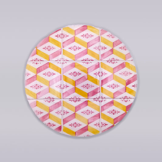 A round Estremoz Ceramic Trivet Hot Pad from Tejo Shop with a geometric pattern featuring interlocking pink, yellow, and white hexagons. Each hexagon contains a smaller pink floral design in the center. The vibrant hand-made hot pad boasts a subtle 3D effect on the pattern, perfect for brightening up kitchen countertops.