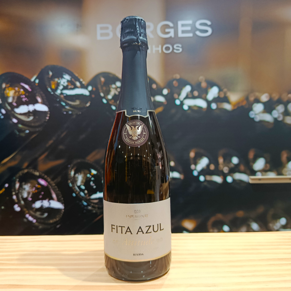 A bottle of Fita Azul Attitude Reserve Brut by Borges Wines stands on a light wooden surface. Blurred wine bottles fill the background, with "BORGES" partially visible. Fine bubbles hint at its refined character.