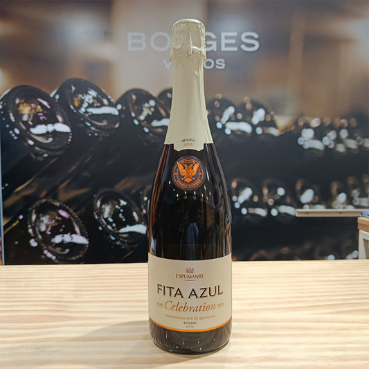 A bottle of Borges Wines Fita Azul Celebration Reserve Medium Dry stands on a wooden surface, its cream label with gold and orange accents reflecting its elegant Portuguese heritage amid blurred rows of wine bottles in the background.