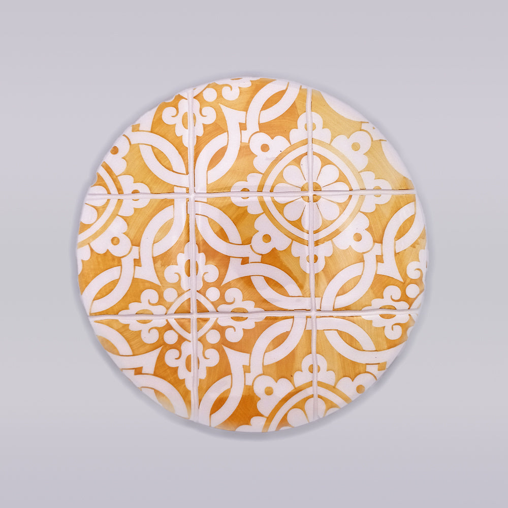 A Guimaraes Ceramic Trivet for Hot Pans from Tejo Shop with an intricate, symmetrical white geometric pattern resembles traditional tile designs. Hand-made with a warm, natural wood background, it brings a touch of artisanal charm to any kitchen. The trivet is set against a plain, grayish backdrop.