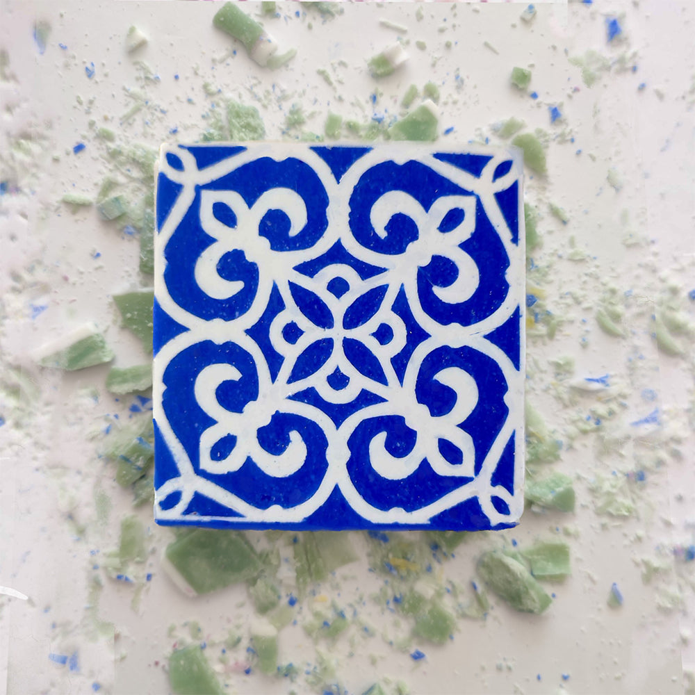 A blue Mosaico Azul Green Tea Soap by Lisbão, featuring a detailed white floral and geometric pattern, rests on a white surface dotted with green and blue fragments, evoking handcrafted Portuguese azulejos.