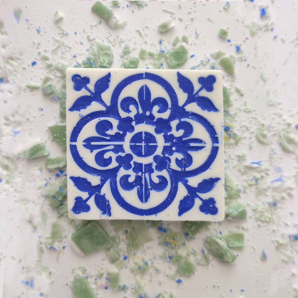 A square Mosaico Azul Green Tea Soap by Lisbão, featuring a blue floral and geometric pattern inspired by Portuguese azulejos, rests on a white surface surrounded by scattered green and blue soap fragments.