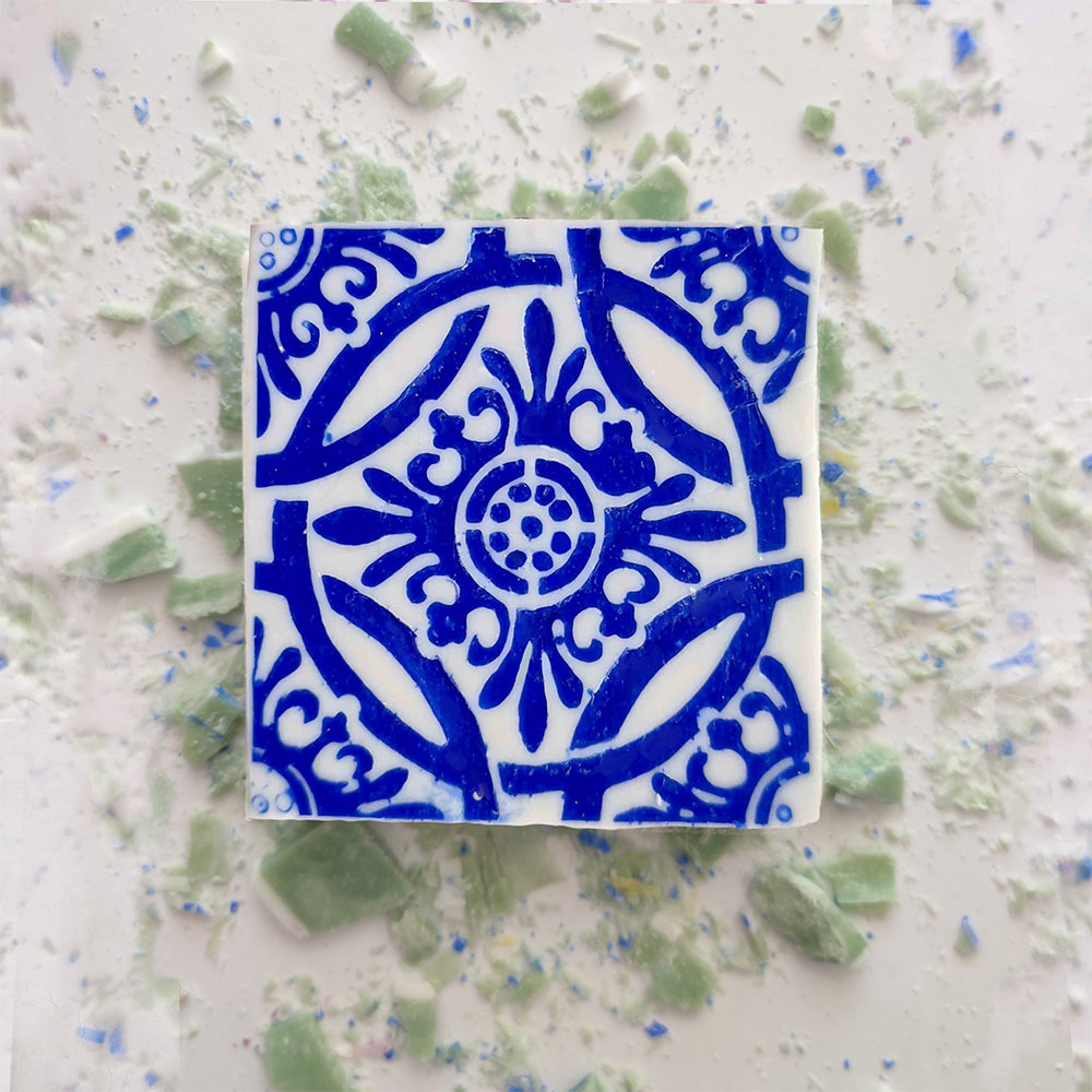 The Mosaico Azul Green Tea Soap by Lisbão features an intricate blue pattern on white, inspired by Portuguese azulejos, and is shown on a white surface with scattered green and blue ceramic fragments.