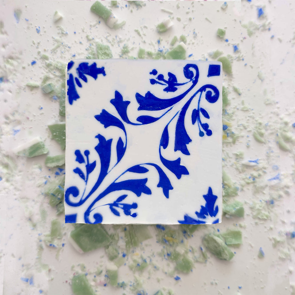 A square ceramic tile with a blue floral pattern sits beside pieces of Lisbão Mosaico Azul Green Tea Soap, handcrafted and inspired by Portuguese azulejos, in green and blue shades.