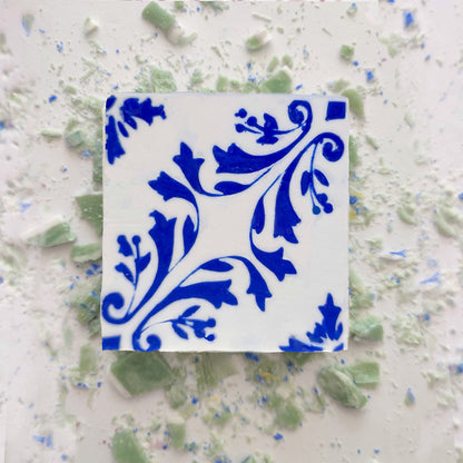 A square ceramic tile with a blue floral pattern sits beside pieces of Lisbão Mosaico Azul Green Tea Soap, handcrafted and inspired by Portuguese azulejos, in green and blue shades.