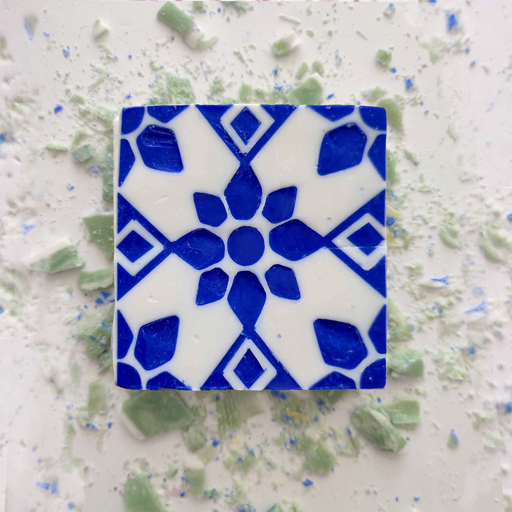 The Lisbão Mosaico Azul Green Tea Soap, shaped like a square tile with a blue and white geometric floral design, rests on scattered light green and blue fragments, evoking handcrafted Portuguese azulejos.