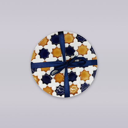 The Heritage Star Mosaic Coaster by Miradouro Ceramics features a Mudéjar star pattern in white, navy blue, and mustard yellow, tied with a dark blue ribbon, displayed on a light gray background.
