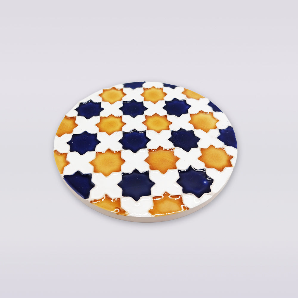 The Heritage Star Mosaic Coaster by Miradouro Ceramics is a round Portuguese ceramic coaster featuring Mudéjar star patterns with navy blue, yellow-orange, and white crosses on a white background.