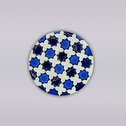 The Heritage Blue Star Mosaic Coaster by Miradouro Ceramics features a symmetrical, azulejo-inspired pattern of interlocking blue and white shapes on a round form, set against a plain light gray background.