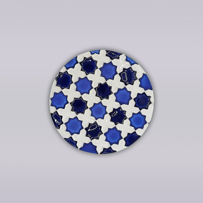 The Heritage Blue Star Mosaic Coaster by Miradouro Ceramics features a round ceramic design with blue and white interlocking stars and crosses, inspired by hand-painted azulejo mosaics, on a light gray background.