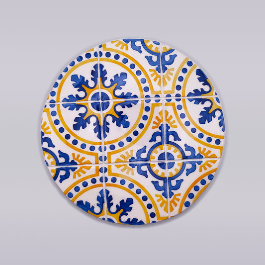 A round ceramic coaster with a vibrant design of blue and yellow intricate patterns resembling traditional Spanish or Moroccan tile work. The intersecting motifs create a symmetrical, eye-catching display against a gray background, making it an essential Island of Madeira Ceramic Tile Trivet by Tejo Shop for any kitchen.