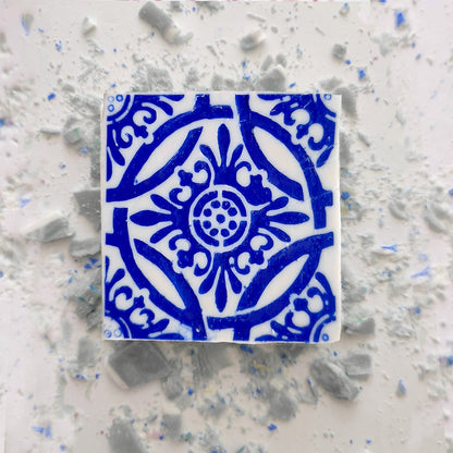 The Jasmine Courtyard Soap by Lisbão sits on a white surface with broken ceramic tiles, echoing the blue floral and geometric artistry of handmade Portuguese tiles.
