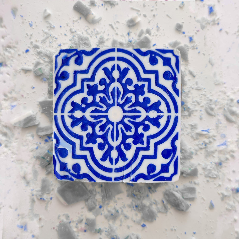 A Lisbão Jasmine Courtyard Soap rests on a light surface beside a decorative blue and white geometric ceramic tile, with scattered dust and broken tile fragments surrounding them.