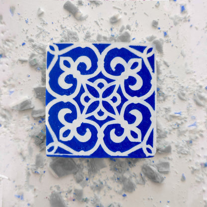 A square blue Jasmine Courtyard Soap by Lisbão, featuring an intricate white floral and geometric pattern, is centered on a pale surface with scattered small gray and blue fragments.