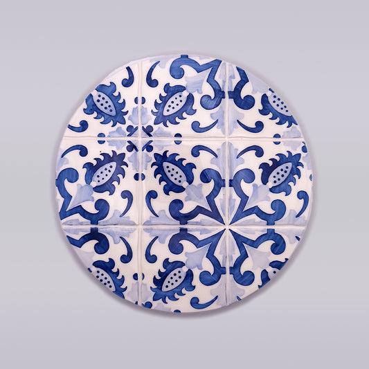 A circular tile design with a pattern of blue and white motifs. The intricate design features floral and geometric shapes, creating a symmetrical and visually appealing arrangement. Perfect for kitchen decor, this Lagos Ceramic Hot Plate Trivet by Tejo Shop has a background with a smooth gradient of light grey.