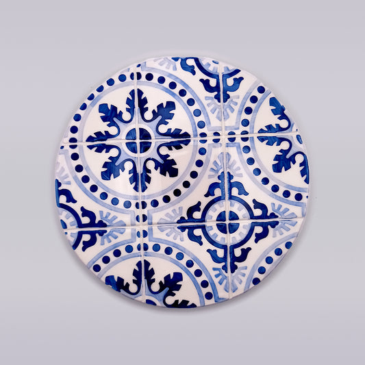A circular arrangement of hand-painted ceramic tiles with intricate blue and white patterns, featuring symmetrical designs and floral motifs, against a plain light gray background. This Liberdade Ceramic Tile Trivet from Tejo Shop brings an air of kitchen luxury to any setting.