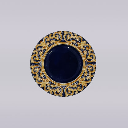 The Manuel I Royal Crest Ceramic Coaster by Miradouro Ceramics features a round shape with a deep blue center and an ornate gold and blue border inspired by Manueline art, set on a light neutral background.