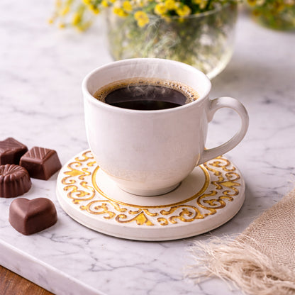 A steaming cup of black coffee rests on the Manueline Golden Crest Ceramic Coaster by Miradouro Ceramics atop a marble surface, with assorted chocolates nearby and a vase of yellow flowers softly blurred in the background.