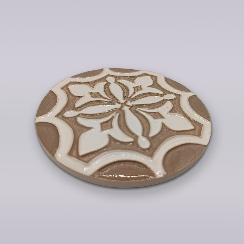 The Ocre Fleur by Miradouro Ceramics is a round hand-painted ceramic coaster featuring a raised geometric and floral design in beige and white, ideal for adding artisanal flair to your décor.