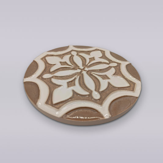 The Ocre Fleur by Miradouro Ceramics is a round hand-painted ceramic coaster featuring a raised geometric and floral design in beige and white, ideal for adding artisanal flair to your décor.
