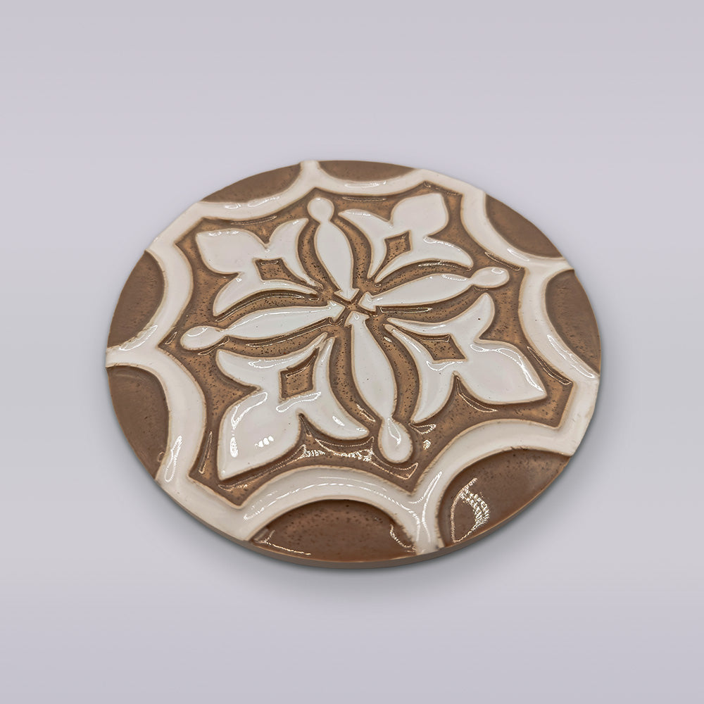 The Ocre Fleur coaster by Miradouro Ceramics is a round, hand-painted brown ceramic piece with a white floral pattern and glossy raised details, bringing artisanal charm to any space.