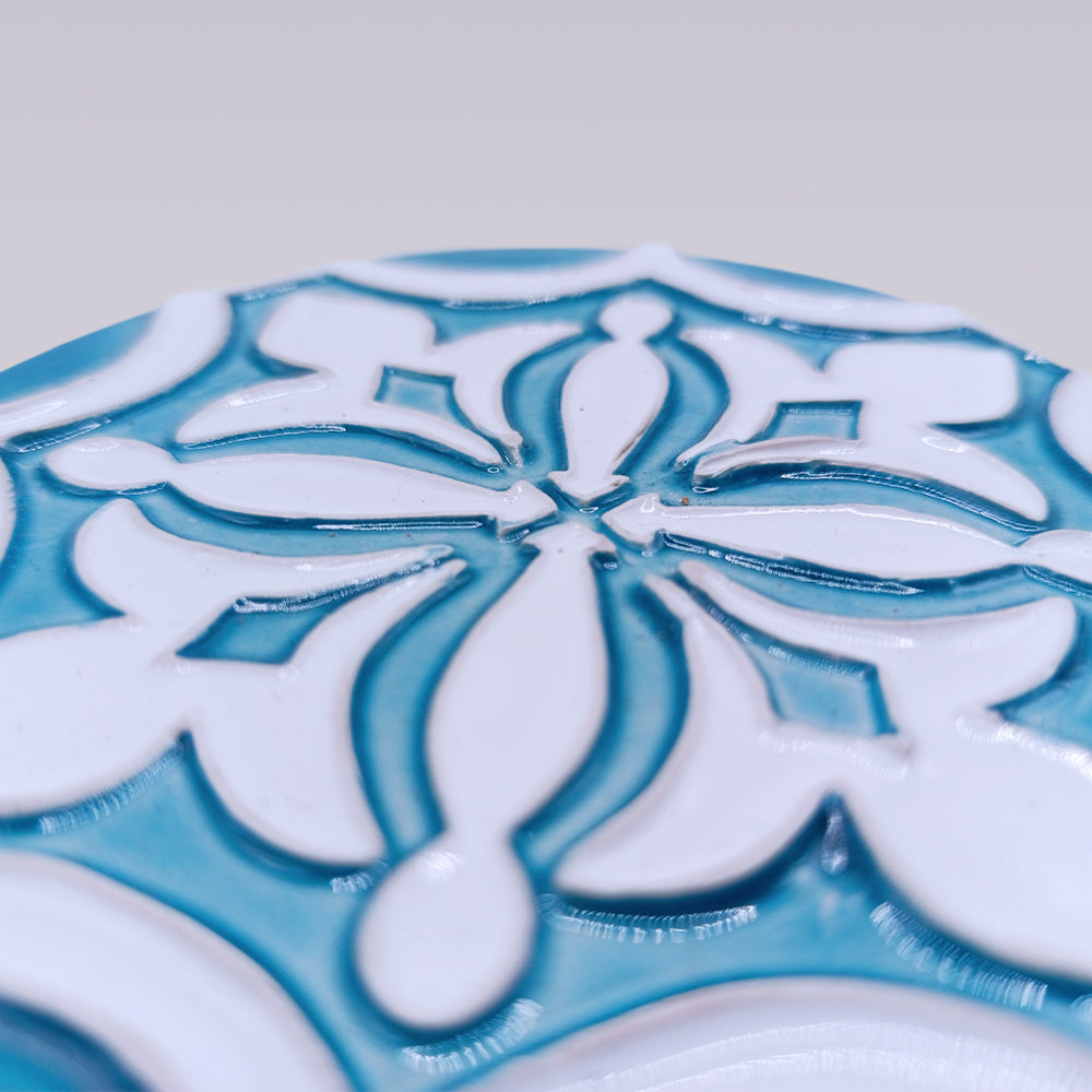 Close-up of the Rosacrux Atlântica ceramic tile by Miradouro Ceramics, showcasing a glossy white and teal blue raised symmetrical floral and geometric pattern on a plain light gray background.